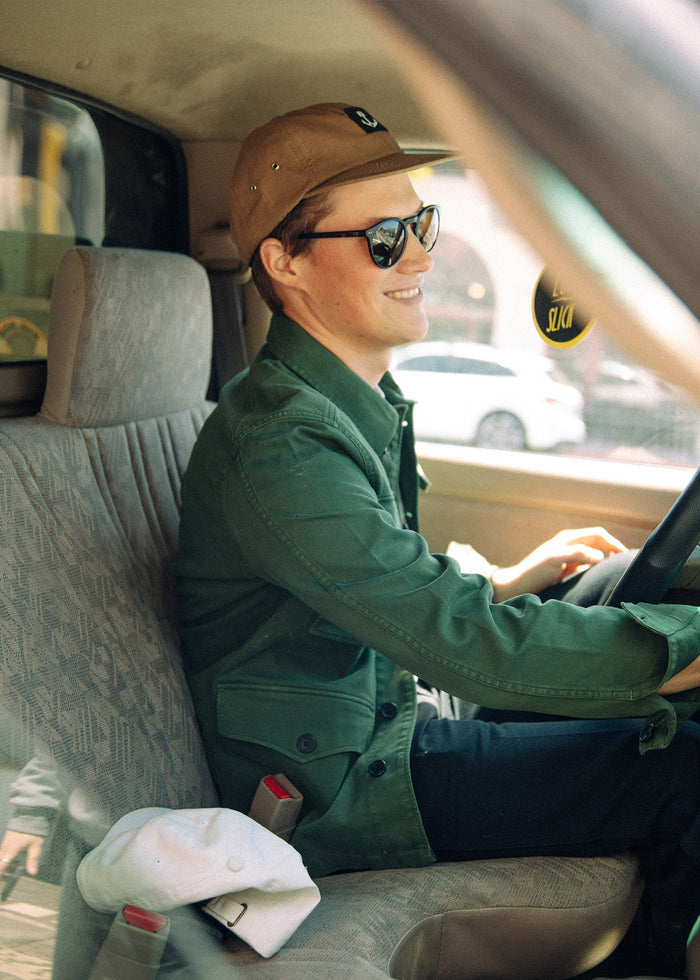 Man in sunglasses and cap smiles while driving a car, wearing a green jacket. A white cap rests on the seat beside him.