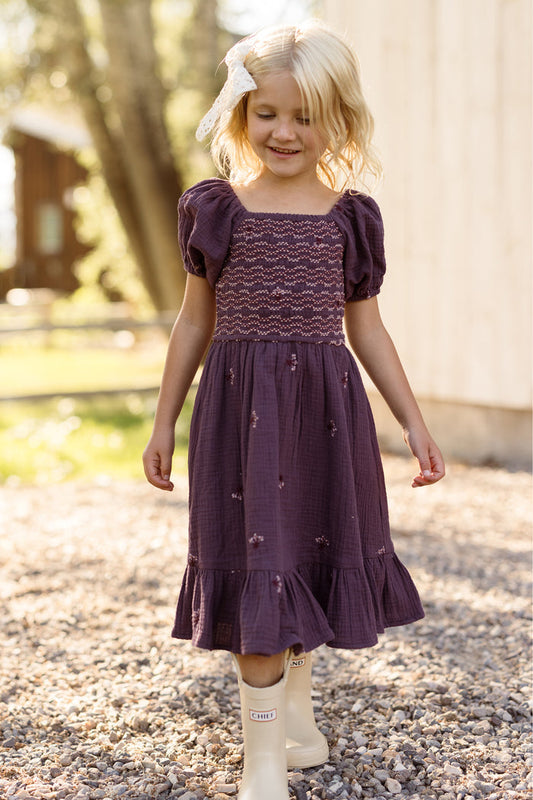 A young girl with blonde hair and a white bow smiles as she walks along a gravel path on a sunny day, wearing the Ivy City Co Mini Acantha Dress in Aubergine Embroidered Gauze with puff sleeves and beige boots.