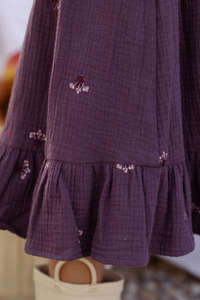 A close-up of the Ivy City Co Mini Acantha Dress in Aubergine Embroidered Gauze reveals its purple textured fabric adorned with delicate, small floral embroidery and a ruffled hem, echoing the intricate charm typical of a cotton smocked dress. Below the dress, part of a cream-colored shoe can be seen.