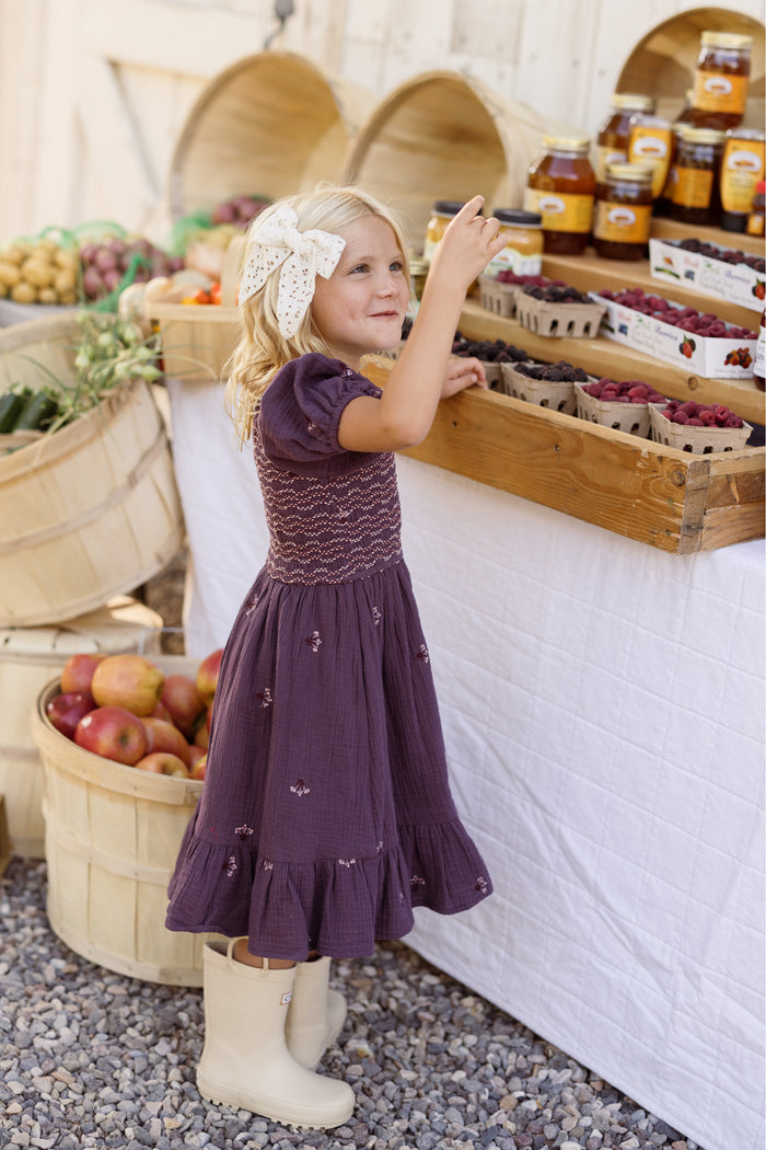 A young girl wearing the Ivy City Co Mini Acantha Dress in Aubergine Embroidered Gauze, featuring puff sleeves, pairs her look with white boots as she selects berries at an outdoor market stand, surrounded by baskets of apples and vegetables and jars of honey on display.