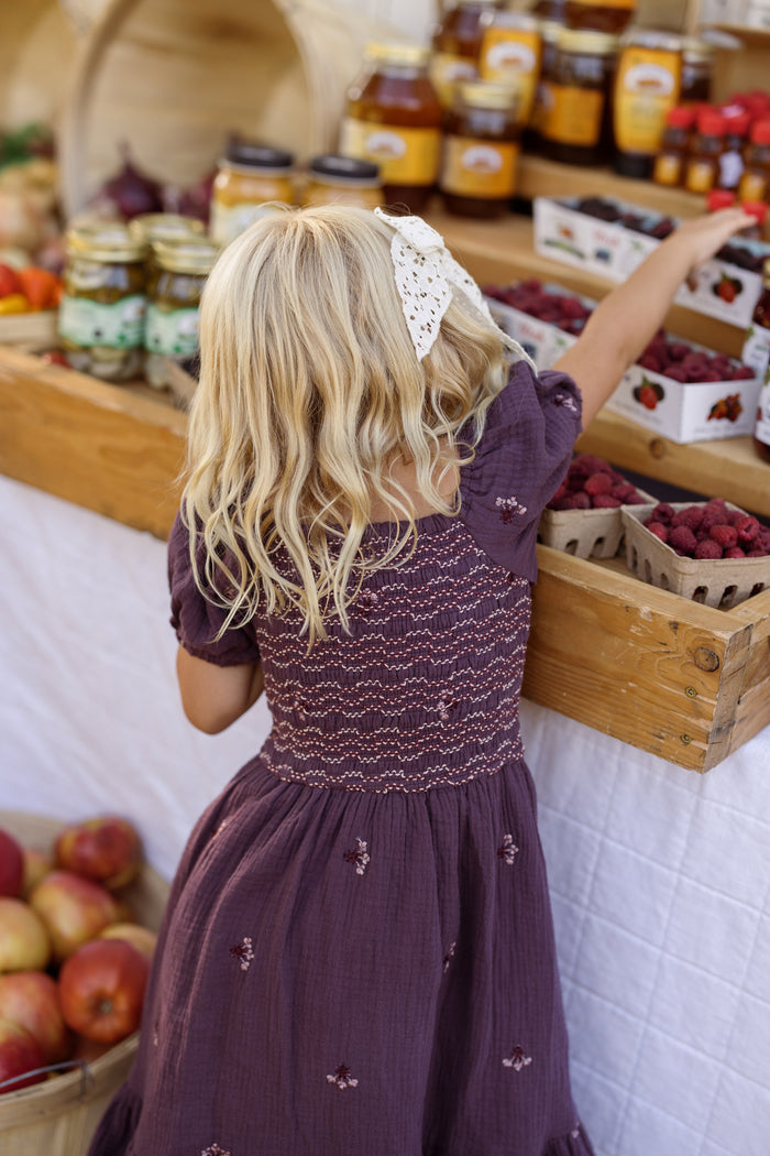 A young girl with blonde hair and a white bow, dressed in the Ivy City Co Mini Acantha Dress in Aubergine Embroidered Gauze—with its charming puff sleeves and smocked detail—reaches for berries at a market stand filled with baskets of fruit, jars of jam, and honey. Apples are visible in the foreground, creating an enchanting scene that highlights this delightful dress.