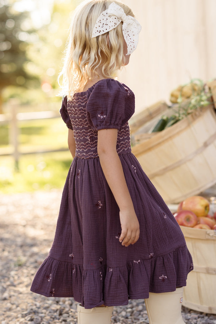 A young girl with blonde hair, wearing a large white bow and the Ivy City Co Mini Acantha Dress in Aubergine Embroidered Gauze with puff sleeves, stands outdoors near baskets of apples on a sunny day.