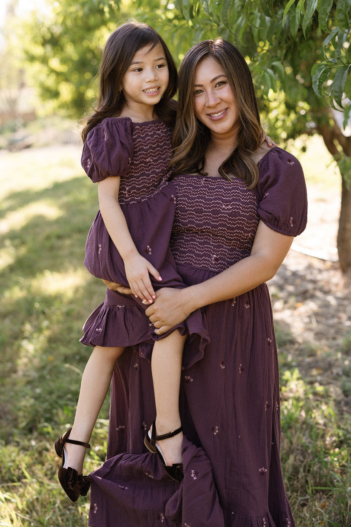 A woman holds a young girl outdoors, both smiling and wearing matching Ivy City Co Mini Acantha Dresses in Aubergine Embroidered Gauze with puff sleeves, standing on grass with trees in the background.