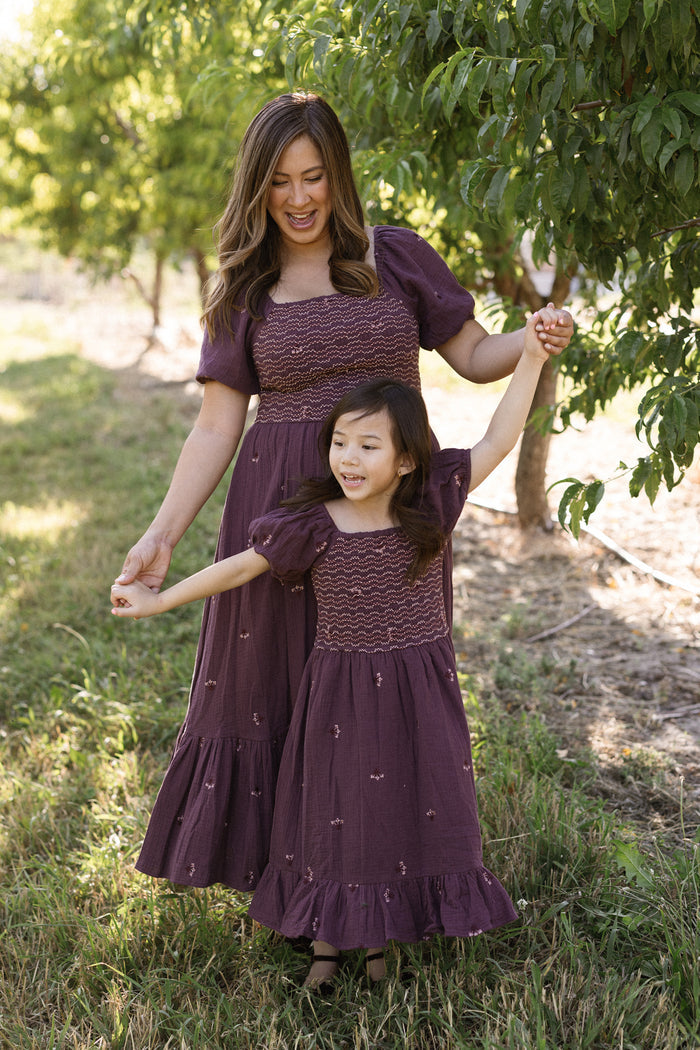 A woman and a young girl, both wearing the Ivy City Co Mini Acantha Dress in Aubergine Embroidered Gauze, smile and hold hands while walking together in a sunny orchard.