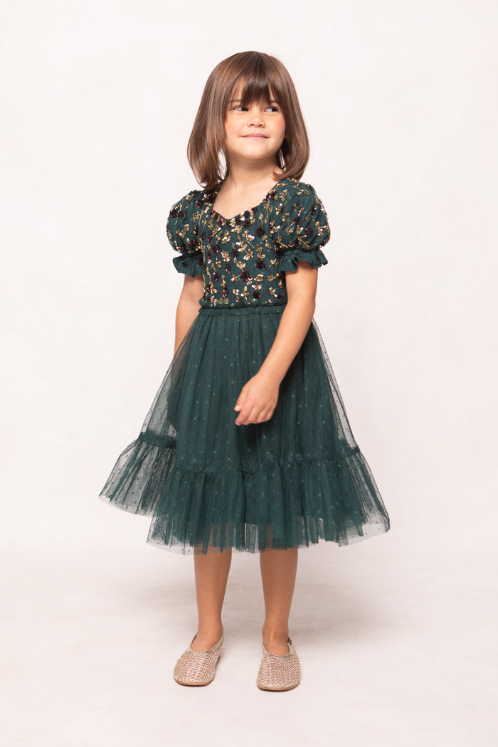A young child with short brown hair smiles softly, wearing Ivy City Co’s Mini Adara Dress in Green—featuring a floral-embroidered bodice and tulle skirt—with beige textured shoes against a plain white background.