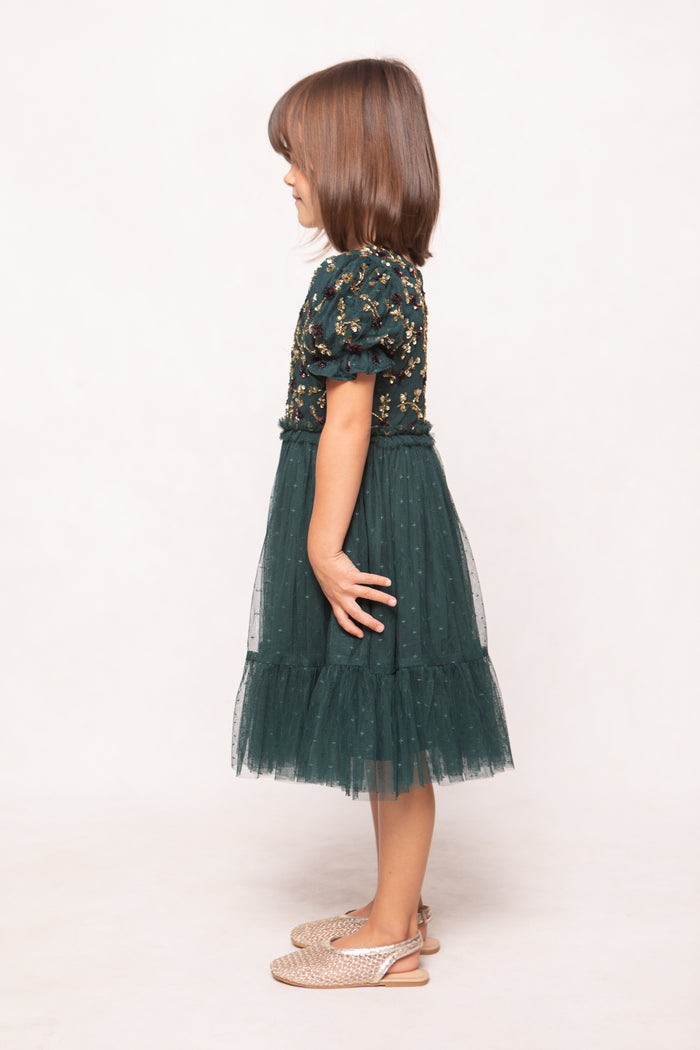 A young girl stands in profile against a plain background, wearing the Ivy City Co Mini Adara Dress in Green, featuring short puff sleeves and an embroidered bodice, paired with woven sandals.