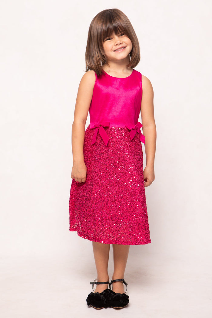 A young girl with brown bobbed hair is smiling and wearing the sleeveless, sparkly Mini Alexis Dress in Fuchsia by Ivy City Co, featuring bow details and a dazzling sequin skirt, paired with black shoes. She stands against a plain white background—perfect for creating a matching look with Mom.