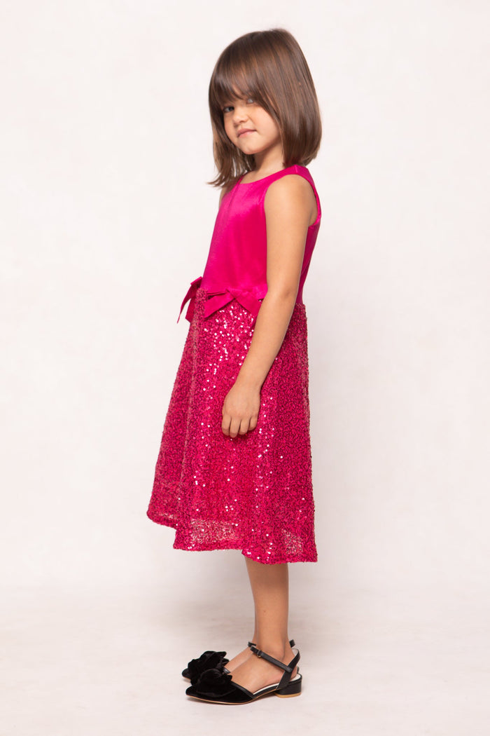 A young girl with straight brown hair stands sideways, wearing the Ivy City Co Mini Alexis Dress in Fuchsia, which features a sleeveless design and a sequin skirt, paired with black sandals against a plain white background—perfect for matching with Mom.