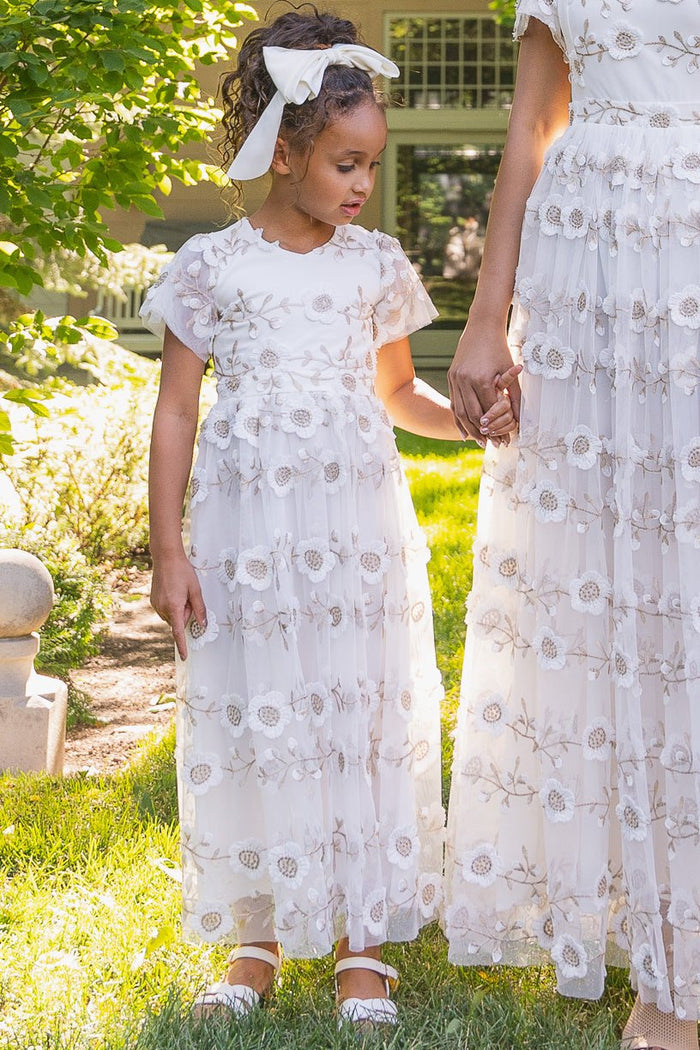 A young girl wears the Ivy City Co Mini Athena Dress in White, holding hands with her mom in matching embroidered outfits, standing together on sunlit grass. The girl's curly hair is tied with a white ribbon and she wears white shoes.