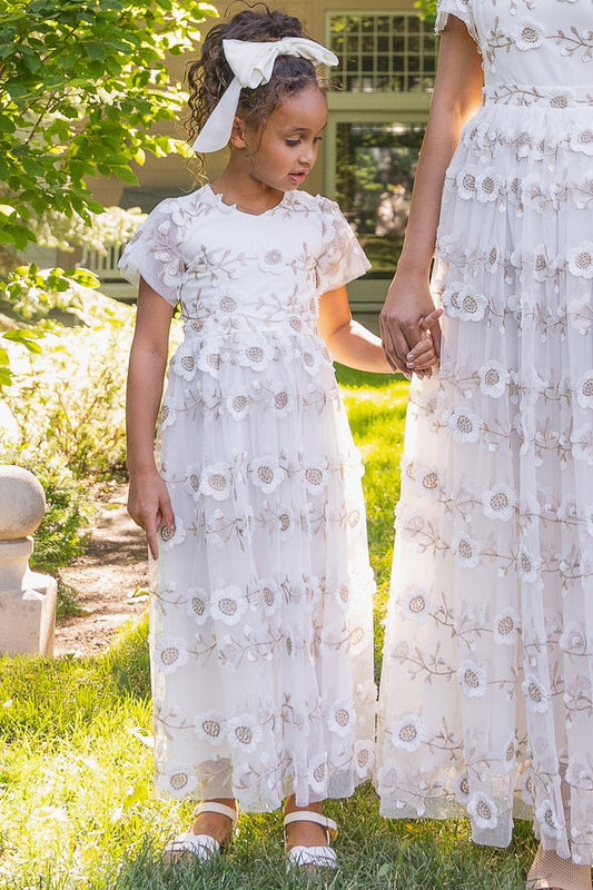 A young girl wears the Ivy City Co Mini Athena Dress in White, holding hands with her mom in matching embroidered outfits, standing together on sunlit grass. The girl's curly hair is tied with a white ribbon and she wears white shoes.