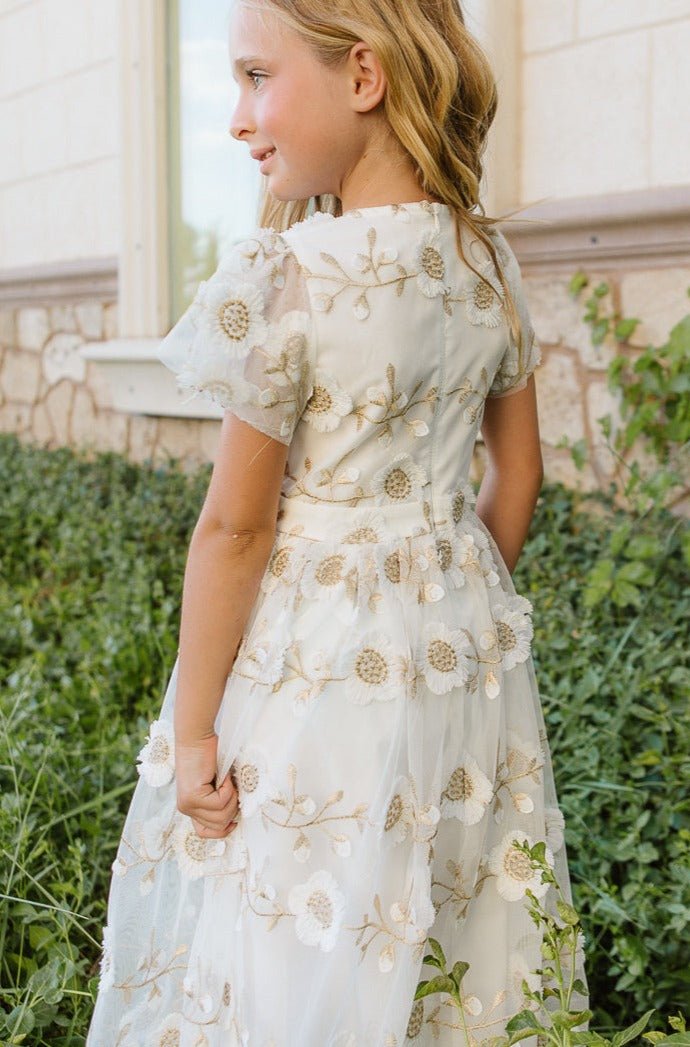 A young girl with blonde hair smiles outdoors in the Ivy City Co Mini Athena Dress in White, featuring floral embroidery and sheer short sleeves—perfect for matching with mom among greenery by a stone building.