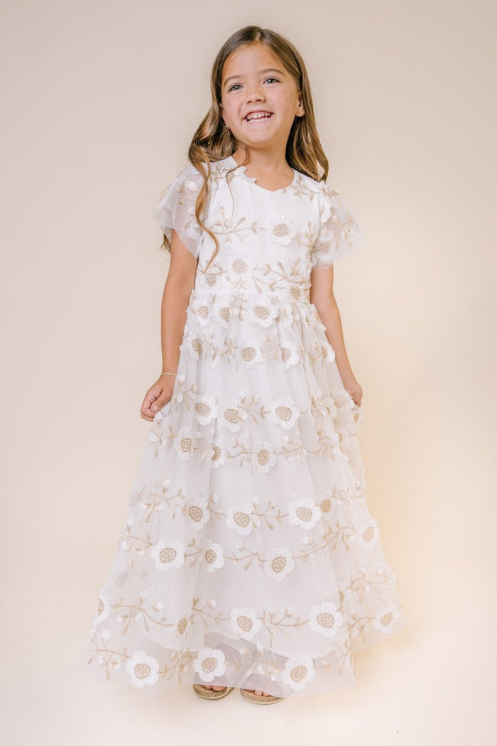 A young girl with long brown hair smiles in the Ivy City Co Mini Athena Dress in White, a flowing embroidered dress with floral details, perfect for matching with mom. She stands against a plain beige background.