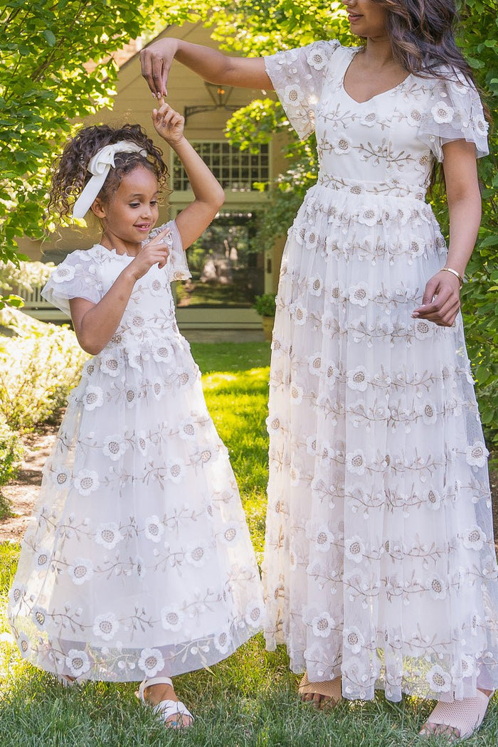 An adult and a child, both dressed in matching Ivy City Co Mini Athena Dresses in White, smile and dance together outside on a sunny day, surrounded by lush greenery.