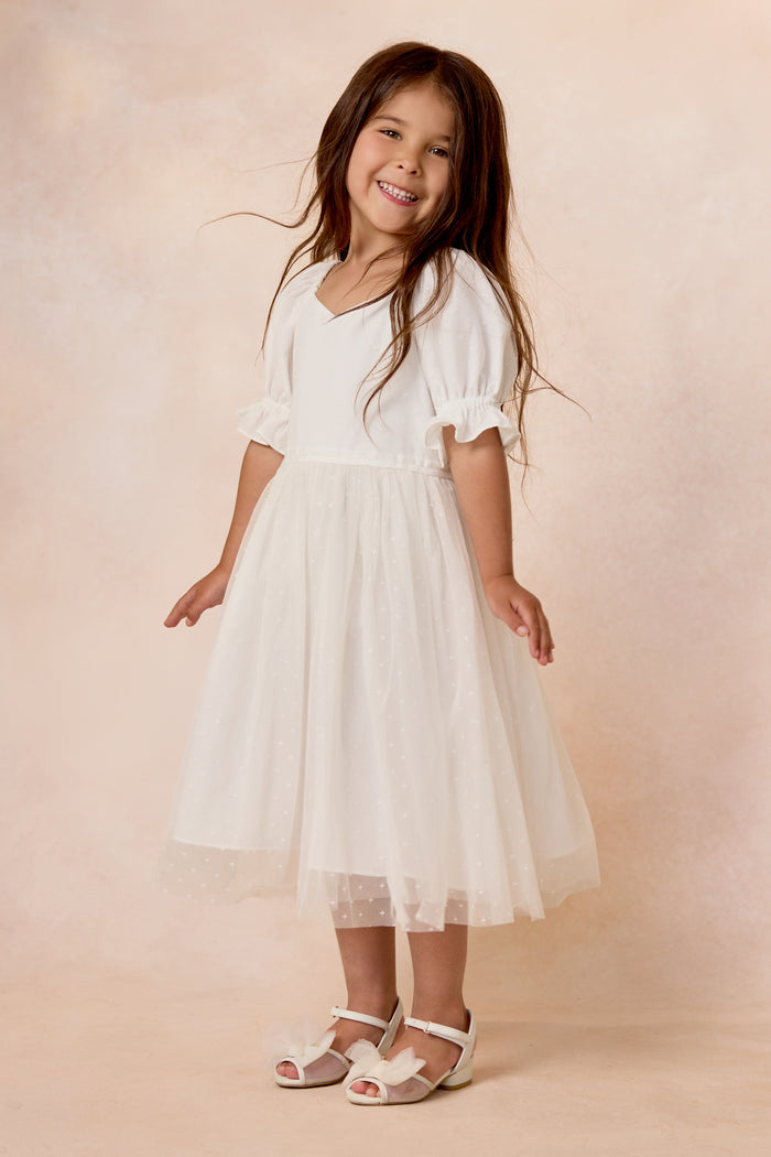 A young girl with long brown hair smiles while wearing the Ivy City Co Mini Ballerina Dress in White and white sandals, standing against a soft beige background.
