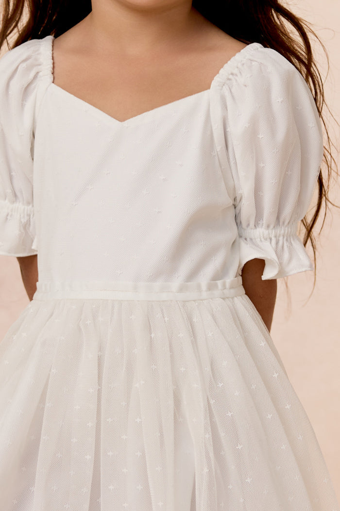 A young girl models the Ivy City Co Mini Ballerina Dress in White, featuring puffed short sleeves and a subtle dotted pattern, standing against a light background with her face not visible.