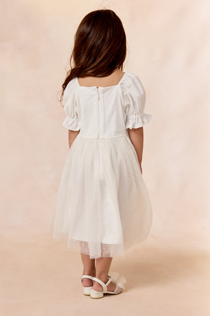 A young girl with long brown hair faces away, wearing the Ivy City Co Mini Ballerina Dress in White with a tulle skirt and white sandals, set against a soft, light background.