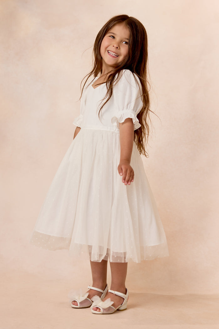 A young girl with long brown hair smiles in the Ivy City Co Mini Ballerina Dress in White, featuring a tulle skirt, paired with white sandals and standing against a soft beige background.