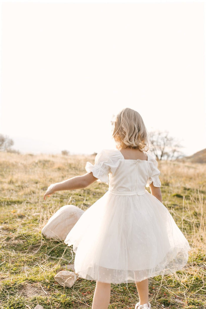 A young child wears the Ivy City Co Mini Ballerina Dress in White, standing in a sunlit grassy field with soft light and distant trees in the background.