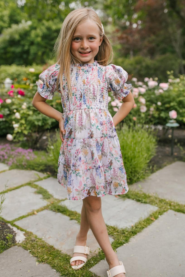 A smiling young girl wears the Ivy City Co Mini Cordelia Floral Dress as she stands on a stone path in a garden, surrounded by blooming flowers and lush greenery.