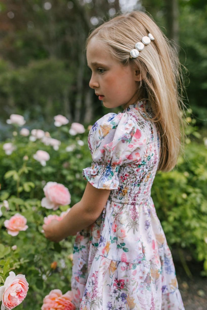 A young girl with long blonde hair, wearing the Ivy City Co Mini Cordelia Floral Dress and a delicate hair clip, stands in a garden surrounded by blooming pink roses, gazing thoughtfully to the side.