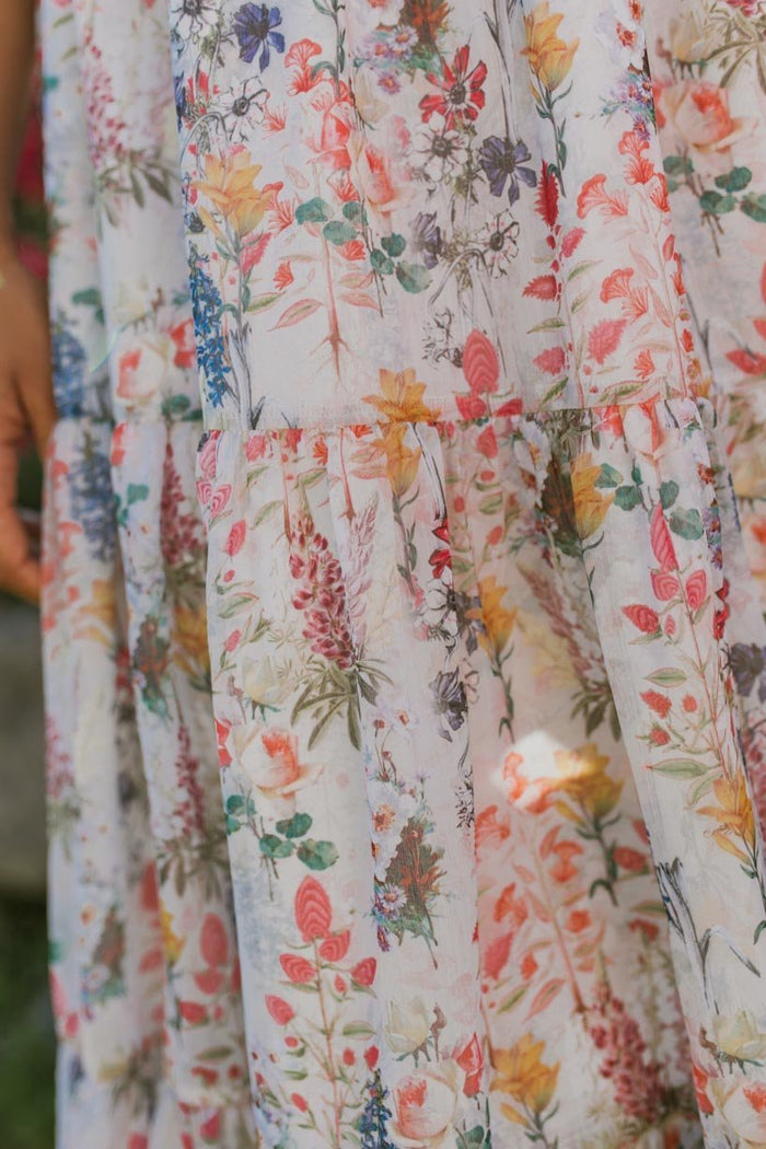 A close-up of the flowing white fabric of the Mini Cordelia Floral Dress by Ivy City Co, adorned with a colorful floral print in shades of pink, yellow, blue, and green. The fabric features gathers and tiered seams, highlighting the charming spring style of this girls’ dress.