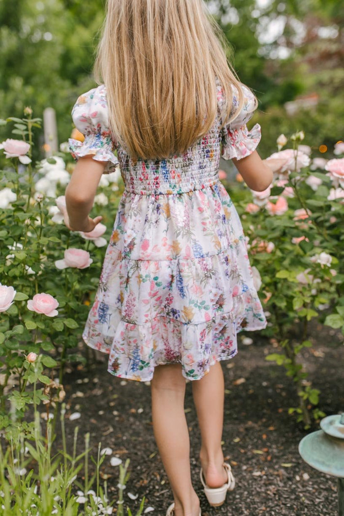 A young girl with long blonde hair, wearing the Ivy City Co Mini Cordelia Floral Dress, walks through a garden filled with blooming pink and white roses.