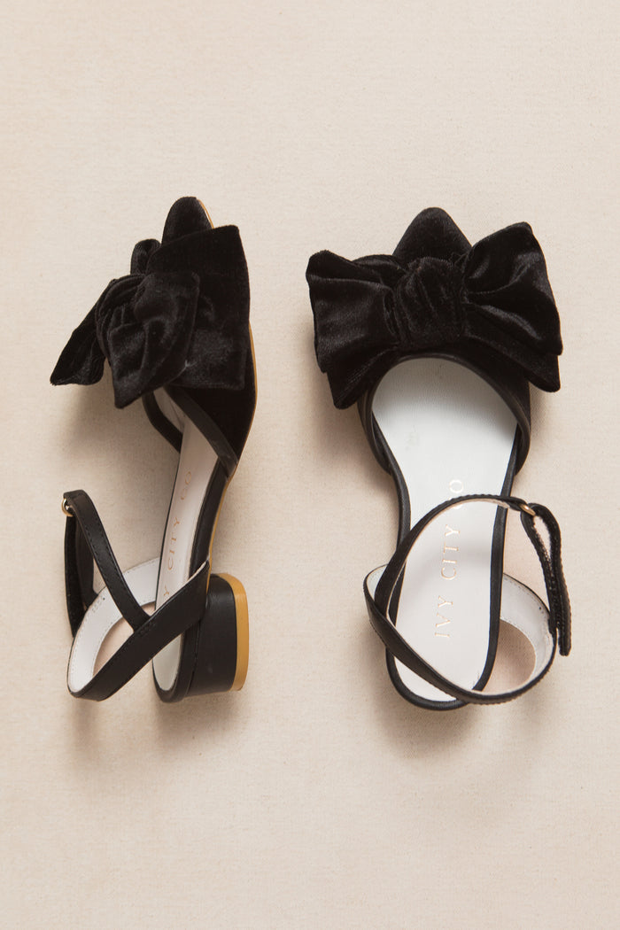 The Mini Eloise Bow Heel in Black by Ivy City Co features open toes, low heels, and large black velvet bows with ankle straps, shown on a light beige background.