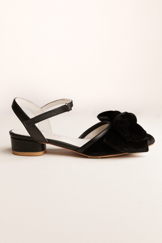 The Mini Eloise Bow Heel in Black by Ivy City Co is a girls' black velvet sandal featuring a low block heel, ankle strap, and large bow—ideal for special occasions. Shown against a plain light background.