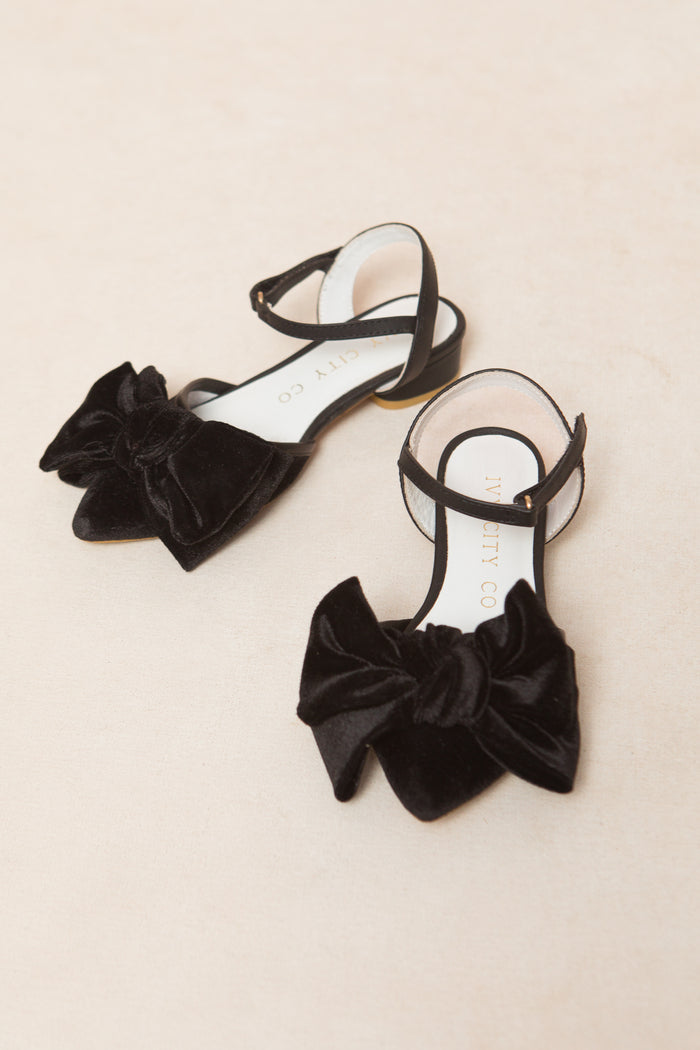 The Mini Eloise Bow Heel in Black by Ivy City Co features black velvet, ankle straps, and large bows on the toes—stylish girls’ shoes displayed on a light beige surface, perfect for any occasion.