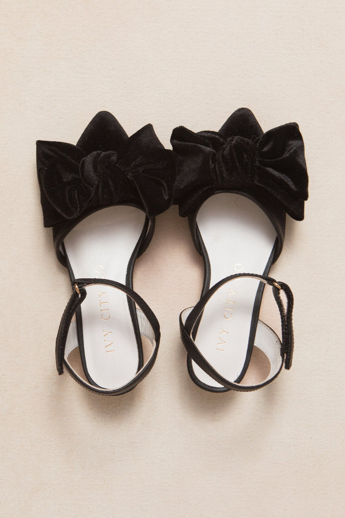The Mini Eloise Bow Heel in Black by Ivy City Co features a pair of black, open-toe sandals with ankle straps and large black velvet bows on the toes, stylishly displayed on a light beige surface.