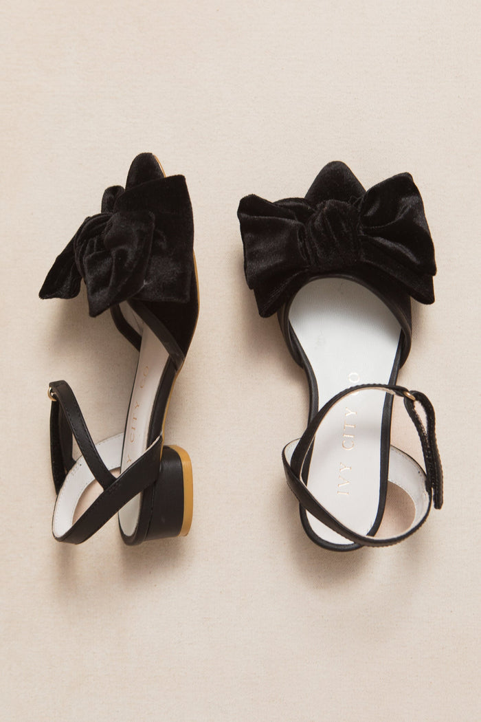 Introducing the Mini Eloise Bow Heel in Black by Ivy City Co — elegant black velvet sandals featuring large bows and ankle straps, beautifully displayed on a beige background. Perfect for anyone seeking sophisticated style with a touch of charm.