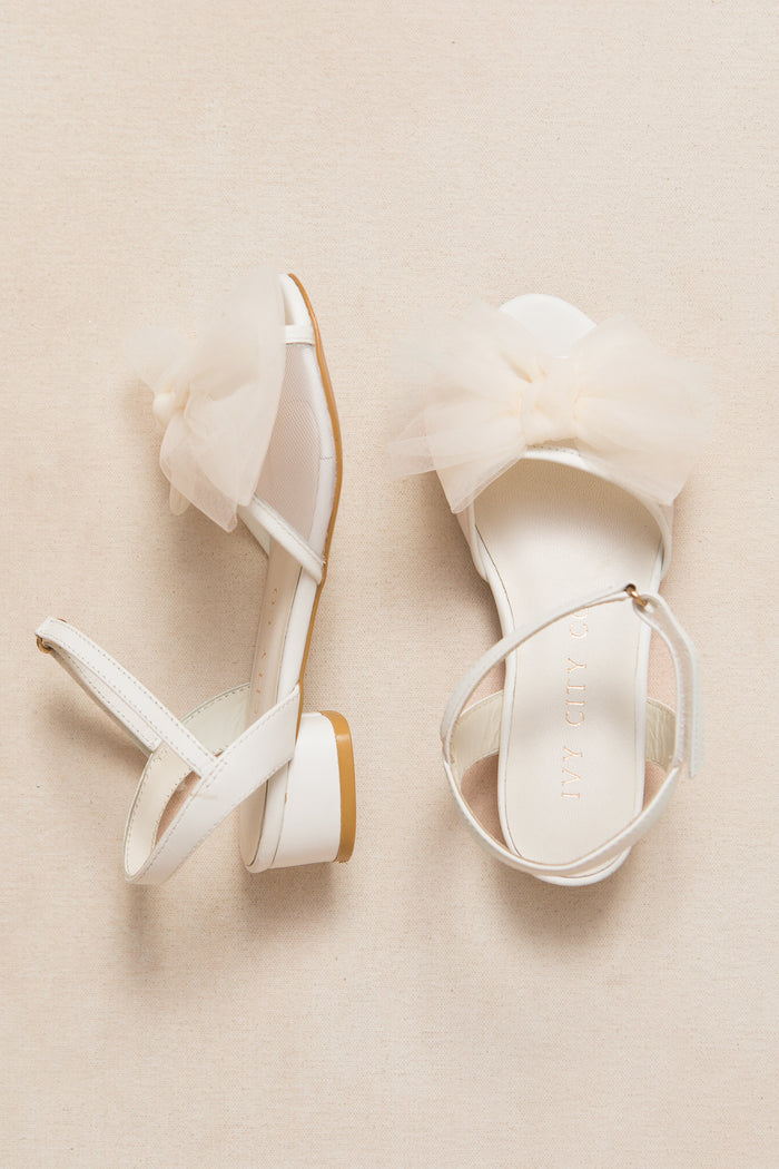 The Mini Ivy Bow Heel in White by Ivy City Co features open toes, low block heels, ankle straps, and oversized sheer tulle bows on the front straps, shown against a neutral beige background.
