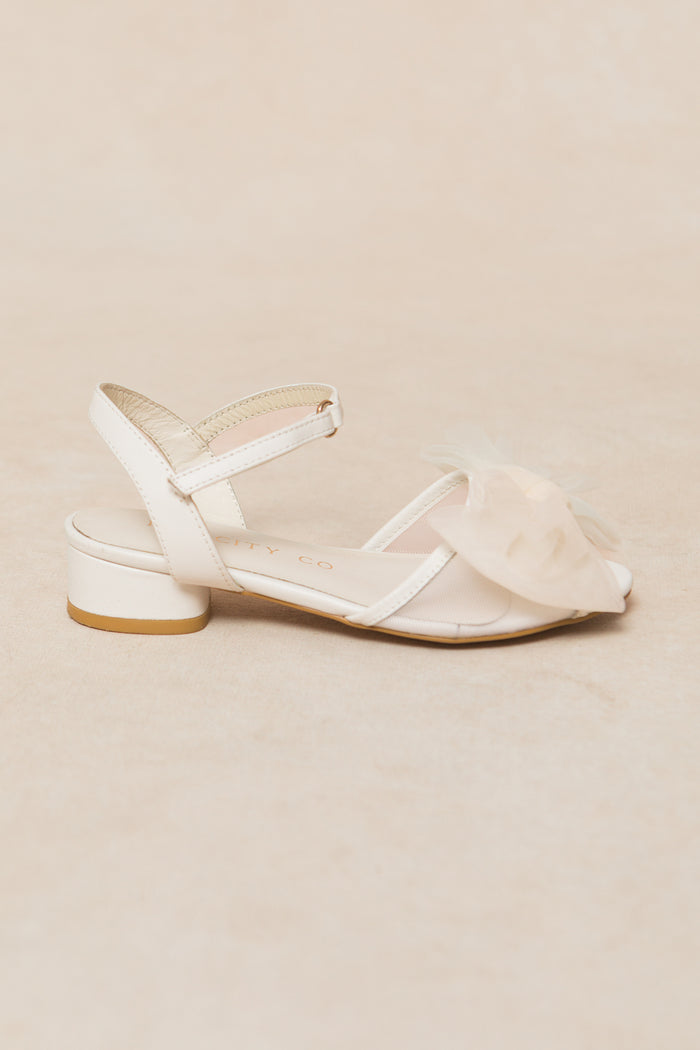 The Ivy City Co Mini Ivy Bow Heel in White, featuring a low block heel, ankle strap, and a large sheer tulle bow on the front, is displayed against a light beige background.