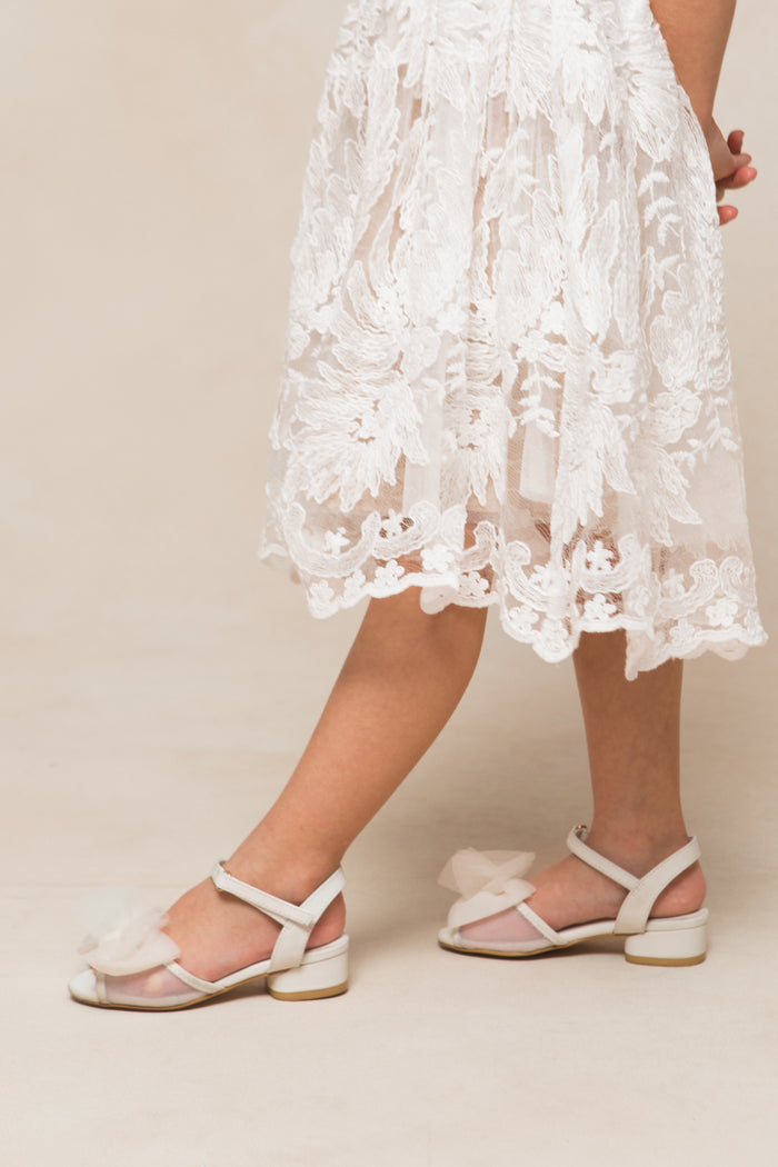 A person wearing a white lace dress and Ivy City Co’s Mini Ivy Bow Heel in White—heels adorned with sheer tulle bows—stands against a neutral background, with only the lower half of their body visible.