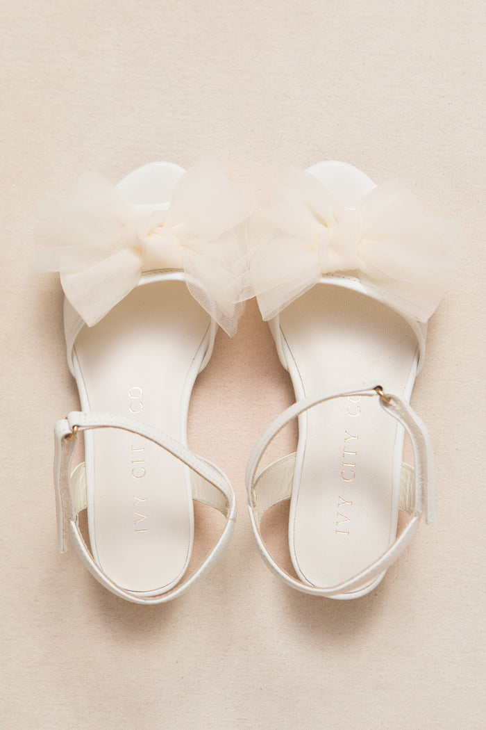 The Mini Ivy Bow Heel in White by Ivy City Co features elegant ankle straps and a large sheer tulle bow on the front, displayed on a light beige surface.