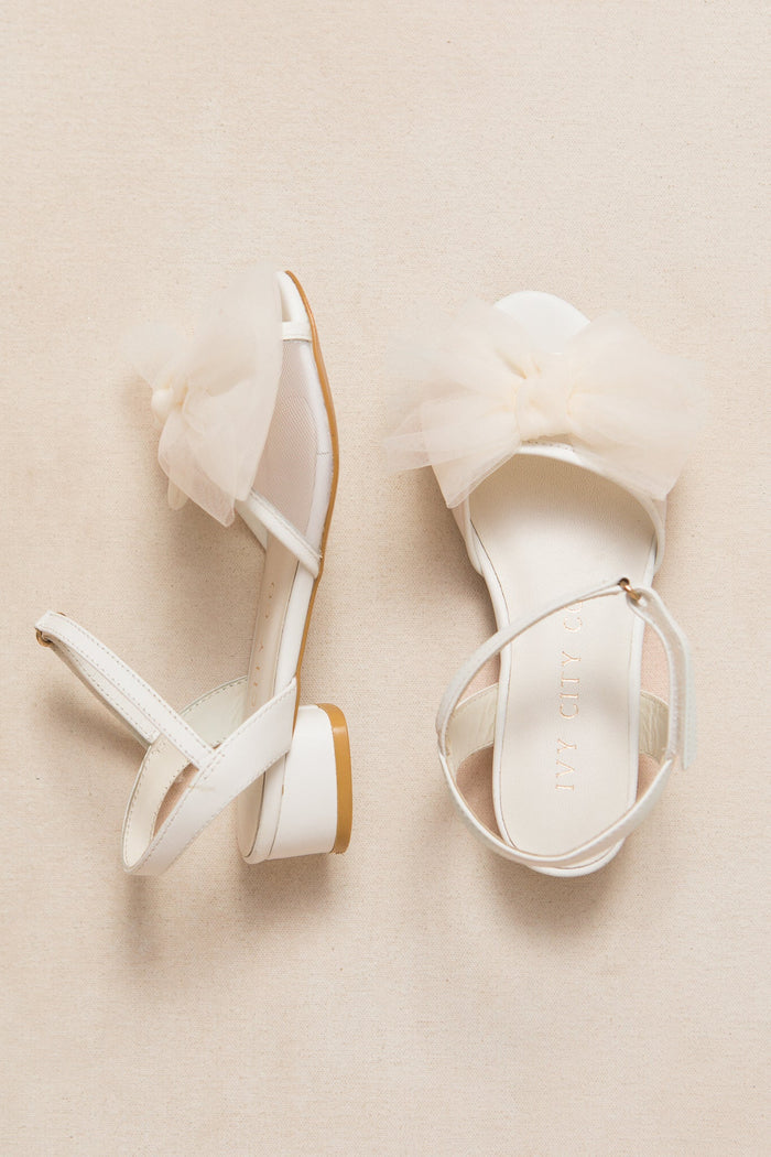 The Mini Ivy Bow Heel in White by Ivy City Co is a pair of white, open-toe sandals with ankle straps and small block heels. Each shoe features a large tulle bow detail on the front for an elegant touch. The comfortable sandals are displayed on a light beige background.
