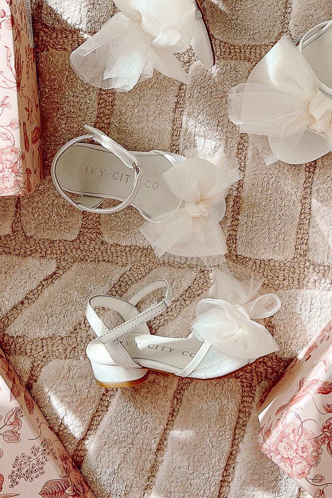The Mini Ivy Bow Heel in White by Ivy City Co, featuring a sheer tulle bow detail and comfortable design, is displayed on a beige patterned rug amidst pink floral-wrapped gift boxes. The insole bears the Ivy City Co label, highlighting its charming style.