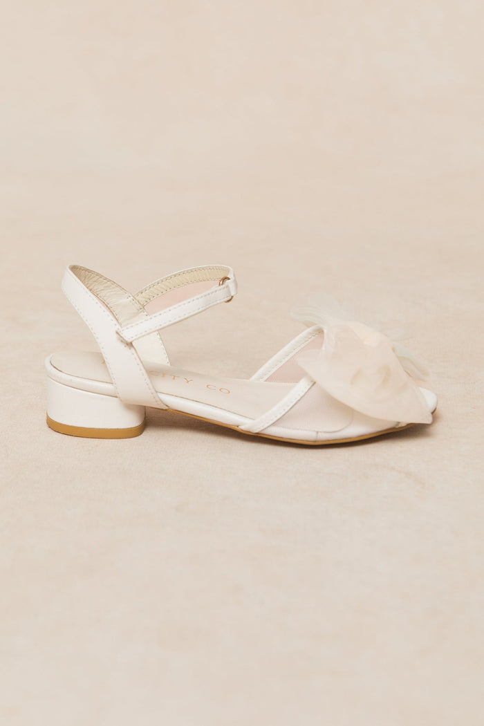 The Mini Ivy Bow Heel in White by Ivy City Co features a single white sandal with a low heel, an ankle strap, and a large tulle bow embellishment on the toe, beautifully displayed on a light beige background—a charming and comfortable addition to your shoe collection.