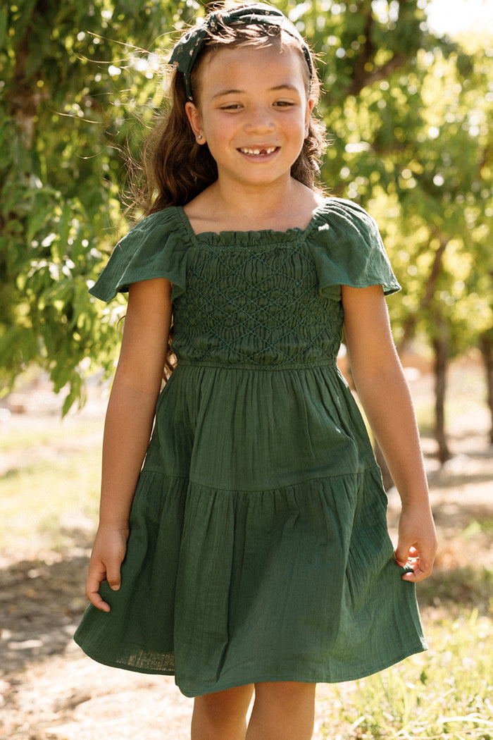 A young girl with long brown hair smiles outdoors among trees and sunlight, wearing the Ivy City Co Mini Jade Dress in Evergreen, which features flutter sleeves and a smocked bodice.