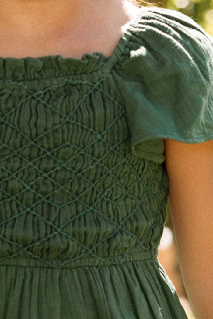 A child wears the Ivy City Co Mini Jade Dress in Evergreen, showcasing a green smocked bodice with delicate puff sleeves. Sunlight illuminates the textured pattern and upper chest, while the background remains softly blurred.