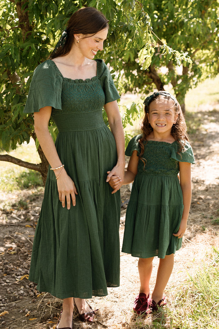 A woman and young girl wear matching dark green smocked dresses with flutter sleeves outdoors near a tree. The woman’s dress is maxi-length, featuring a beautifully detailed smocked bodice; the girl wears the Ivy City Co Mini Jade Dress in Evergreen, which is knee-length and echoes the same charming style.