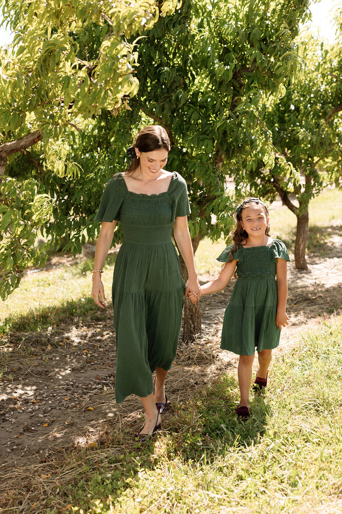 A woman and a young girl, both wearing matching Ivy City Co Mini Jade Dresses in Evergreen featuring flutter sleeves and a smocked bodice, hold hands and smile as they walk beneath leafy trees in a sunlit orchard.