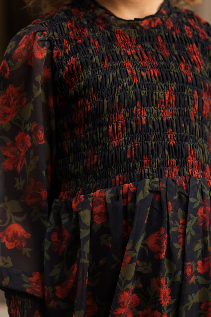 A person wearing the Ivy City Co Mini Linden Dress in Scarlet Blooms, featuring red floral patterns with green leaves, smocking on the bodice, and sheer long balloon sleeves.