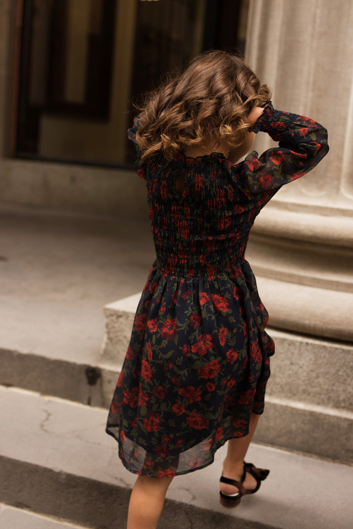 A young girl with curly hair, wearing the Ivy City Co Mini Linden Dress in Scarlet Blooms, walks up stone steps with her hands in her hair. The background features a large column and a building entrance.