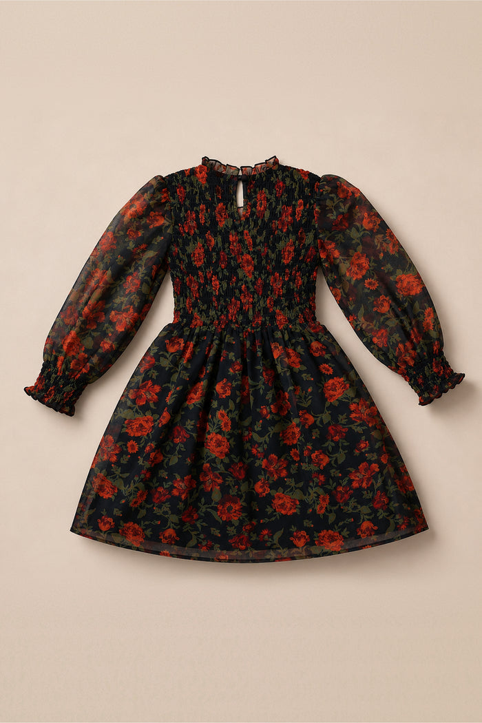 The Mini Linden Dress in Scarlet Blooms by Ivy City Co is a charming girls’ dress featuring a black long-sleeve silhouette adorned with red and green floral patterns. Designed with a smocked bodice, subtle ruffled cuffs, and a delicate ruffle at the collar, this balloon sleeve dress is elegantly displayed flat on a beige background.
