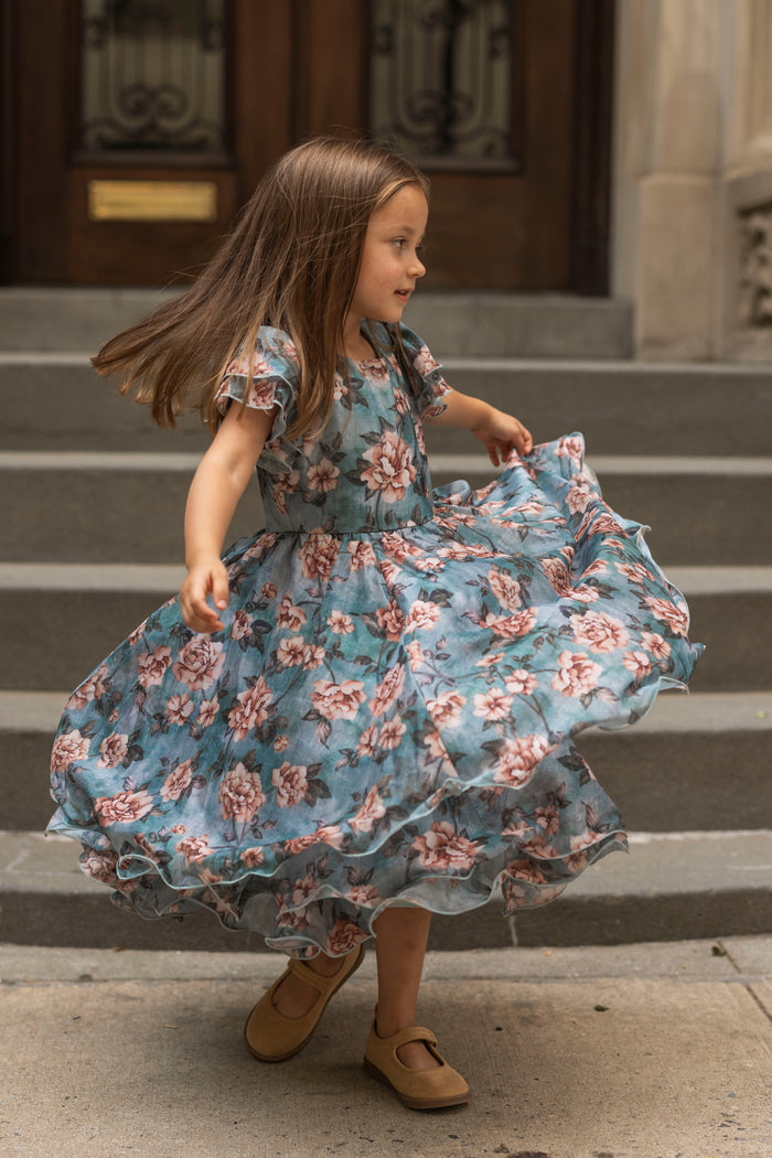 A young girl in the Ivy City Co Mini Lisette Dress in Rose Dusk spins joyfully on a stone sidewalk before the steps, her hair and skirt fanning out as she smiles, capturing a lighthearted elegance that’s perfect for a junior bridesmaid dress at a fall wedding.