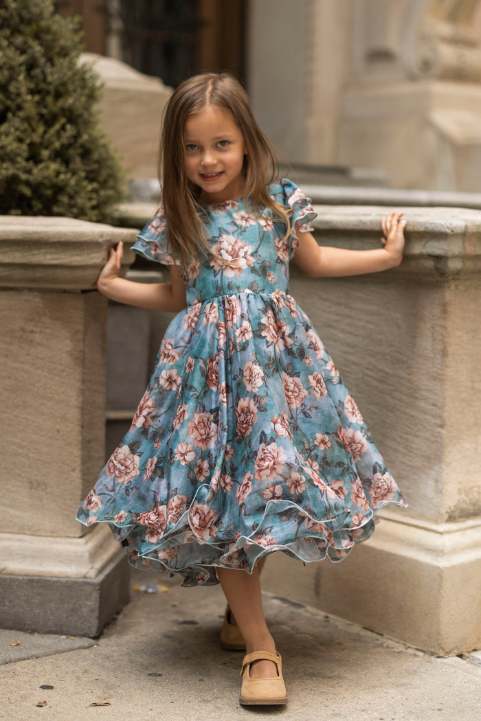 A young girl in the Ivy City Co Mini Lisette Dress in Rose Dusk and tan shoes smiles while posing outdoors, leaning with both hands on a stone railing—the perfect style for a junior bridesmaid dress at a fall wedding.