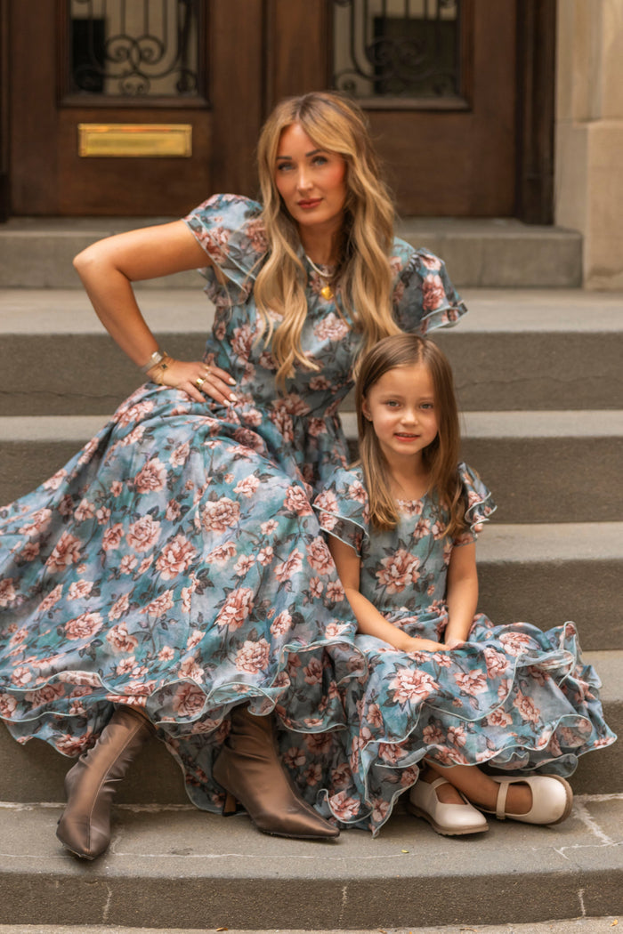 A woman and a young girl sit on stone steps, both wearing matching Ivy City Co Mini Lisette Dresses in Rose Dusk. The woman, with long blonde hair and boots, sits beside the girl—whose brown hair and white shoes make her look perfect for a junior bridesmaid dress at a fall wedding. They both face the camera.
