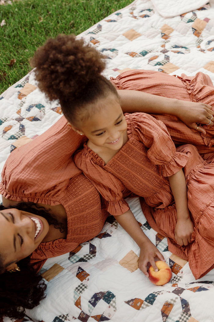 A woman and a young girl, both wearing matching Ivy City Co Mini Madeline Dresses in Apple Cider with puff sleeves, relax on a patterned picnic blanket. The girl smiles while holding a peach, and they both appear happy, enjoying time together outdoors.