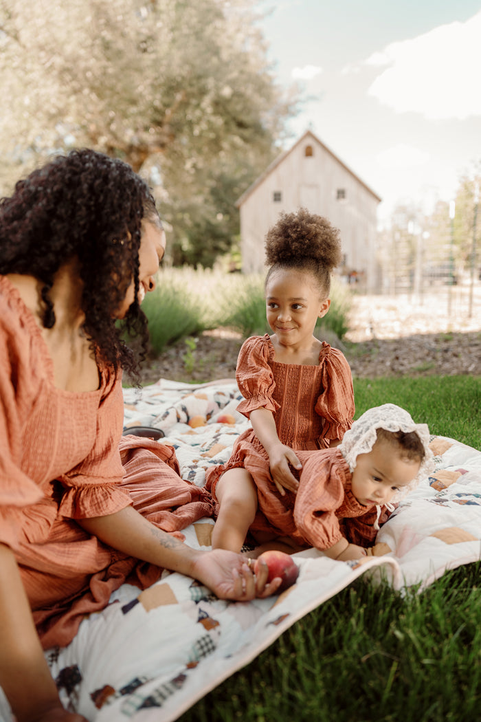 A woman and two young children sit on a blanket outdoors, all wearing matching rust-colored outfits from Ivy City Co. The children are dressed in the Mini Madeline Dress in Apple Cider, featuring charming puff sleeves and a smocked bodice for a sweet touch. One child smiles while sitting up, while the other crawls on the blanket, with a barn and trees providing a picturesque backdrop.